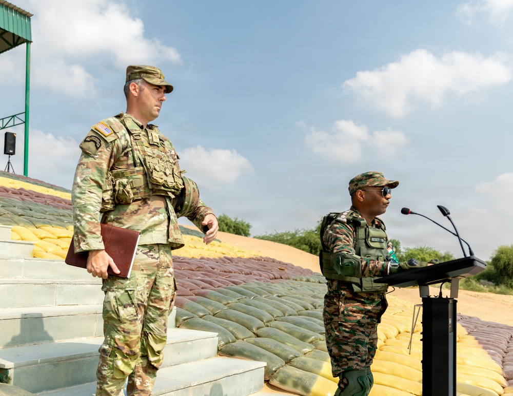 Two military personnel in camouflage uniforms are positioned at what appears to be an outdoor military ceremony or briefing. One soldier, wearing US Army OCP (Operational Camouflage Pattern) uniform with a tactical vest and patrol cap, stands on steps holding a folder. A second soldier in a different camouflage pattern stands at a podium equipped with microphones, suggesting an official address or ceremony.