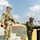 Indian Army leader delivers remarks during Yudh Abhyas 2026 Two military personnel in camouflage uniforms are positioned at what appears to be an outdoor military ceremony or briefing. One soldier, wearing US Army OCP (Operational Camouflage Pattern) uniform with a tactical vest and patrol cap, stands on steps holding a folder. A second soldier in a different camouflage pattern stands at a podium equipped with microphones, suggesting an official address or ceremony.