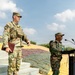 Two military personnel in camouflage uniforms are positioned at what appears to be an outdoor military ceremony or briefing. One soldier, wearing US Army OCP (Operational Camouflage Pattern) uniform with a tactical vest and patrol cap, stands on steps holding a folder. A second soldier in a different camouflage pattern stands at a podium equipped with microphones, suggesting an official address or ceremony.