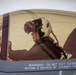 A military pilot is seated inside the cockpit of a fighter jet, visible through the tinted canopy. The pilot is wearing a flight helmet with a raised visor and appears to be in a pre-flight or ground operations phase. A warning label on the canopy frame reads 'WARNING - DO NOT CUT CANOPY WITHIN 3 INCHES OF CANOPY FRAME.'