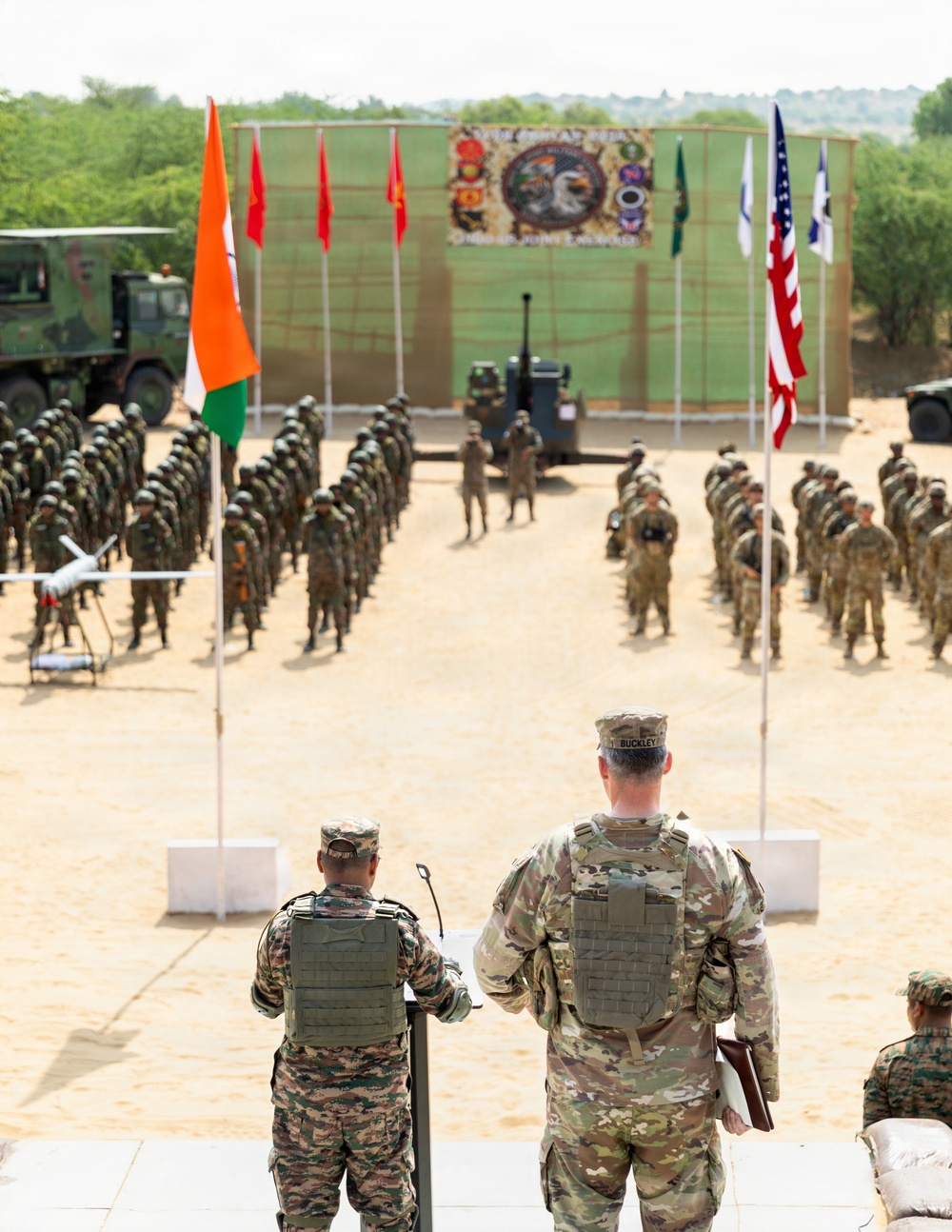 A joint military ceremony or parade is taking place on an open sandy ground, with two formations of soldiers standing at attention on either side of a central aisle. Two senior officers — one in Indian Army camouflage and one in US Army MultiCam uniform with the name tape 'BUCKLEY' — are observing from an elevated platform. Flags of India and the United States are prominently displayed, along with a ceremonial backdrop banner suggesting a bilateral military exercise.