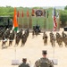 A joint military ceremony or parade is taking place on an open sandy ground, with two formations of soldiers standing at attention on either side of a central aisle. Two senior officers — one in Indian Army camouflage and one in US Army MultiCam uniform with the name tape 'BUCKLEY' — are observing from an elevated platform. Flags of India and the United States are prominently displayed, along with a ceremonial backdrop banner suggesting a bilateral military exercise.