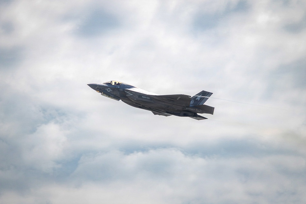 A U.S. Marines F-35B stealth fighter jet is captured in flight against a partly cloudy sky. The aircraft bears visible markings including 'MARINES' text, a unit designation that appears to read 'VMFA-533', and the number '5' on the tail. The jet is banking slightly, providing a clear view of its underside and stealth-optimized airframe.