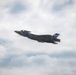 A U.S. Marines F-35B stealth fighter jet is captured in flight against a partly cloudy sky. The aircraft bears visible markings including 'MARINES' text, a unit designation that appears to read 'VMFA-533', and the number '5' on the tail. The jet is banking slightly, providing a clear view of its underside and stealth-optimized airframe.