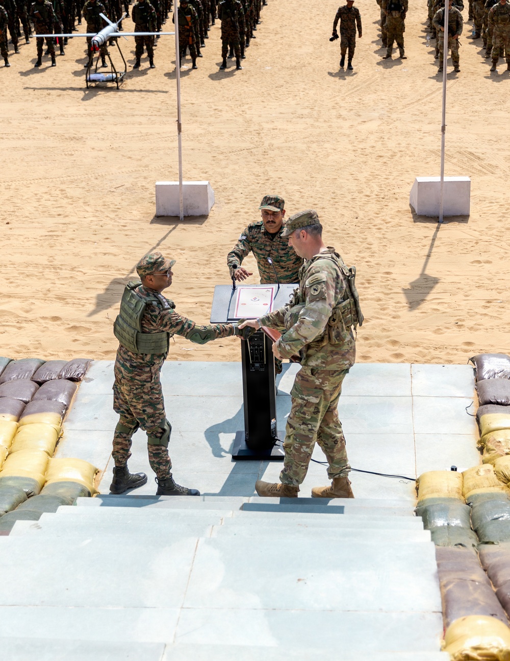 A military ceremony is taking place on an elevated platform in a desert/sandy environment. Two soldiers in different camouflage uniforms are exchanging or presenting a framed document/certificate at a podium, while a third soldier observes. A large formation of troops is visible in the background along with what appears to be a UAV/drone on display.