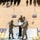 U.S., Indian Army leaders strengthened partnership at Yudh Abhas 2026 A military ceremony is taking place on an elevated platform in a desert/sandy environment. Two soldiers in different camouflage uniforms are exchanging or presenting a framed document/certificate at a podium, while a third soldier observes. A large formation of troops is visible in the background along with what appears to be a UAV/drone on display.