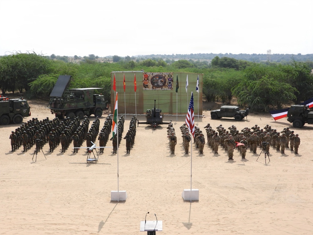 A military joint exercise ceremony is taking place on an open sandy parade ground, with two large formations of soldiers from what appear to be multiple nations standing at attention or saluting. National flags including the Indian tricolor, the United States flag, and what appears to be a French flag are prominently displayed on poles. Military vehicles including what appears to be a missile launcher system and artillery pieces are positioned in the background alongside a ceremonial backdrop board.
