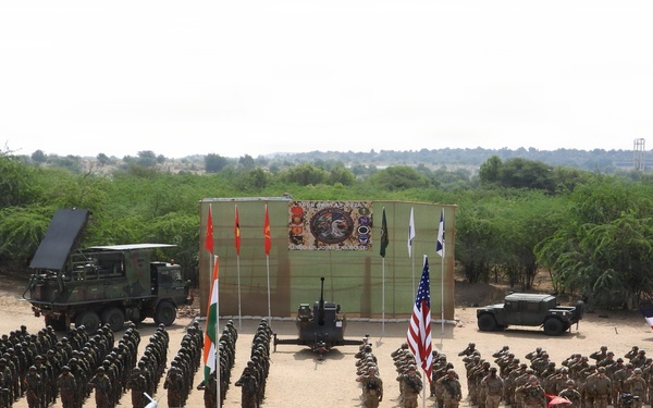 A military joint exercise ceremony is taking place on an open sandy parade ground, with two large formations of soldiers from what appear to be multiple nations standing at attention or saluting. National flags including the Indian tricolor, the United States flag, and what appears to be a French flag are prominently displayed on poles. Military vehicles including what appears to be a missile launcher system and artillery pieces are positioned in the background alongside a ceremonial backdrop board.