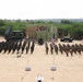 A military joint exercise ceremony is taking place on an open sandy parade ground, with two large formations of soldiers from what appear to be multiple nations standing at attention or saluting. National flags including the Indian tricolor, the United States flag, and what appears to be a French flag are prominently displayed on poles. Military vehicles including what appears to be a missile launcher system and artillery pieces are positioned in the background alongside a ceremonial backdrop board.