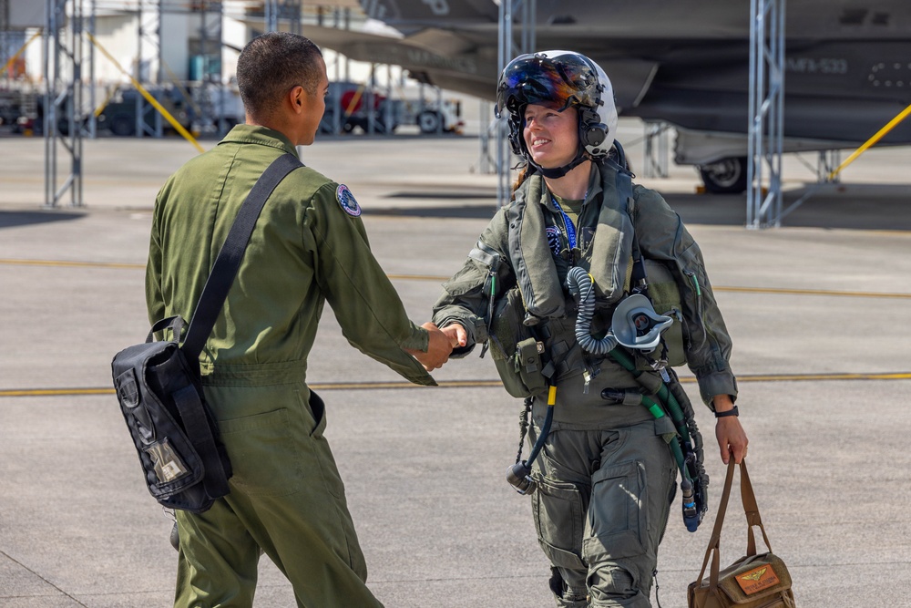 Two military aviators in olive drab flight suits are shaking hands on an aircraft flight line or tarmac. The pilot on the right is fully equipped with a flight helmet, oxygen mask, G-suit, survival vest, and is carrying a flight bag, suggesting they have just returned from or are preparing for a flight. Military jet aircraft, likely F-35s, are visible in the background inside a hangar or on the ramp.