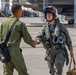 Two military aviators in olive drab flight suits are shaking hands on an aircraft flight line or tarmac. The pilot on the right is fully equipped with a flight helmet, oxygen mask, G-suit, survival vest, and is carrying a flight bag, suggesting they have just returned from or are preparing for a flight. Military jet aircraft, likely F-35s, are visible in the background inside a hangar or on the ramp.
