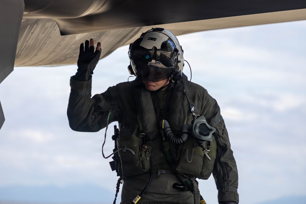 A military aviator in full flight gear is conducting a pre-flight or post-flight inspection of an aircraft, touching the underside of the fuselage or wing. The individual is equipped with a flight helmet, oxygen mask, survival vest, and olive drab flight suit. The image is taken from a low angle against an overcast sky, giving a dramatic perspective.