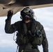 A military aviator in full flight gear is conducting a pre-flight or post-flight inspection of an aircraft, touching the underside of the fuselage or wing. The individual is equipped with a flight helmet, oxygen mask, survival vest, and olive drab flight suit. The image is taken from a low angle against an overcast sky, giving a dramatic perspective.