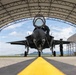 A fifth-generation stealth fighter jet, consistent with an F-35 Lightning II, is positioned inside an arched military hangar shelter on a flight line. Ground crew personnel in flight suits are visible, with one appearing to assist a pilot in the open cockpit and another standing to the right of the aircraft. Mountains and a partly cloudy sky are visible in the background through the hangar opening.