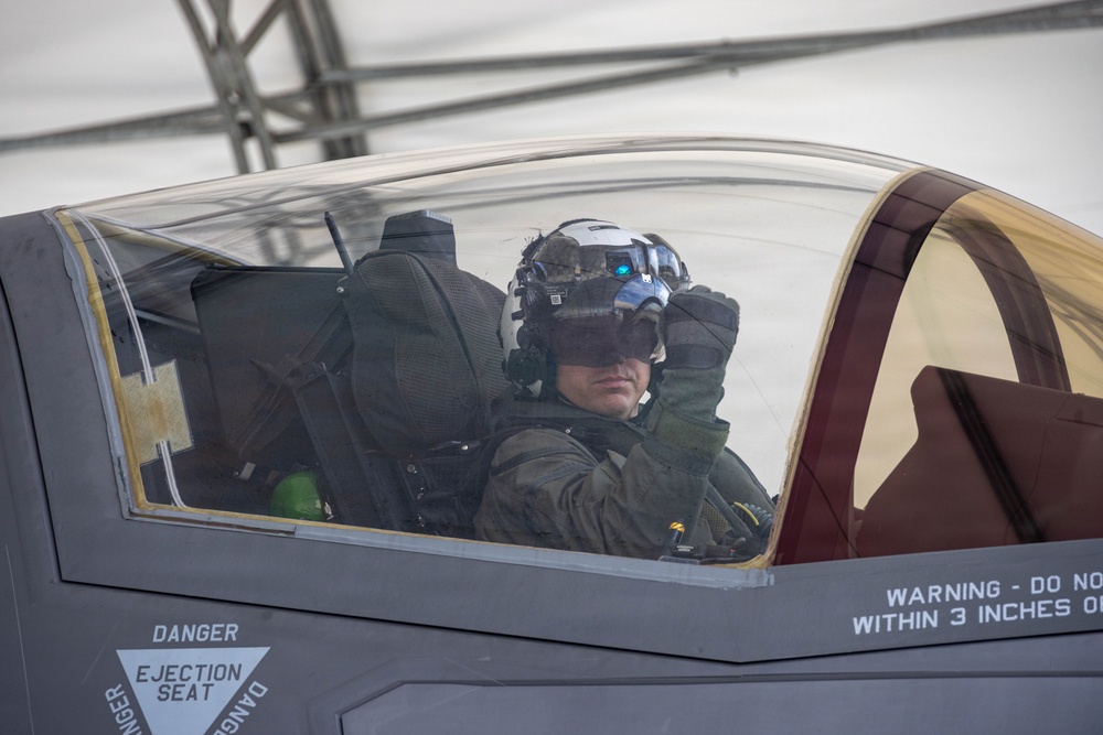 A military pilot is seated in the cockpit of what appears to be an F-35 Lightning II fighter jet, wearing a helmet-mounted display system with visible blue optical sensors. The canopy is open and the pilot appears to be conducting pre-flight checks or preparing for a mission. Warning labels including 'DANGER - EJECTION SEAT' and 'WARNING - DO NOT WITHIN 3 INCHES O[F]' are clearly visible on the aircraft fuselage.