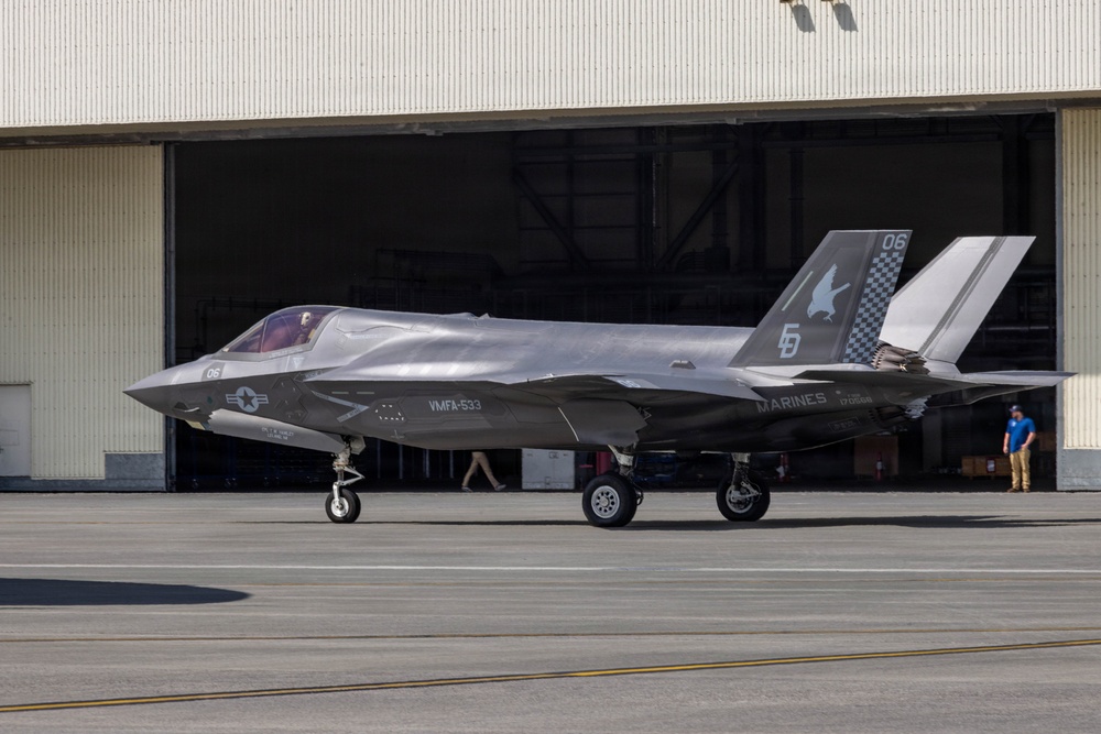 A U.S. Marines F-35B stealth fighter jet is parked on a tarmac in front of a large military hangar. The aircraft bears the markings of VMFA-533 squadron with tail number '06' and distinctive squadron artwork on the vertical stabilizer. Two personnel are visible near the hangar in the background.