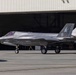 A U.S. Marines F-35B stealth fighter jet is parked on a tarmac in front of a large military hangar. The aircraft bears the markings of VMFA-533 squadron with tail number '06' and distinctive squadron artwork on the vertical stabilizer. Two personnel are visible near the hangar in the background.