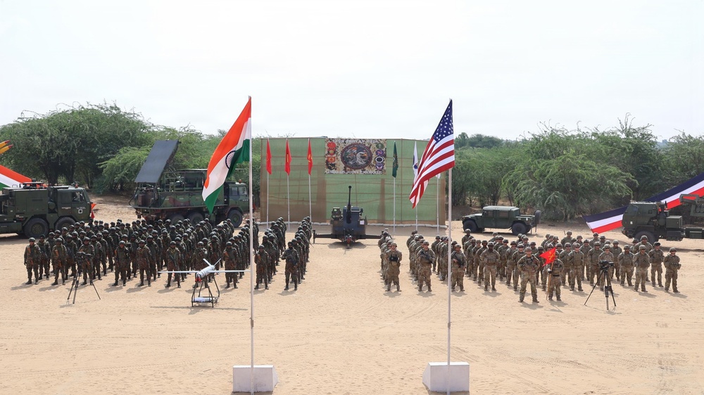 A joint military exercise or ceremony is taking place on an open sandy ground, featuring soldiers from multiple nations standing in formation. Two prominent flags — the Indian tricolor and the United States flag — are displayed on poles in the foreground, with additional red flags and what appears to be a partially visible flag on the right edge. Military vehicles, weapon systems, and a drone are also displayed as part of the event.