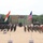 U.S., Indian Armies stand together at Yudh Abhas 2026 A joint military exercise or ceremony is taking place on an open sandy ground, featuring soldiers from multiple nations standing in formation. Two prominent flags — the Indian tricolor and the United States flag — are displayed on poles in the foreground, with additional red flags and what appears to be a partially visible flag on the right edge. Military vehicles, weapon systems, and a drone are also displayed as part of the event.