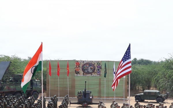 A joint military exercise or ceremony is taking place on an open sandy ground, featuring soldiers from multiple nations standing in formation. Two prominent flags — the Indian tricolor and the United States flag — are displayed on poles in the foreground, with additional red flags and what appears to be a partially visible flag on the right edge. Military vehicles, weapon systems, and a drone are also displayed as part of the event.