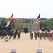 A joint military exercise or ceremony is taking place on an open sandy ground, featuring soldiers from multiple nations standing in formation. Two prominent flags — the Indian tricolor and the United States flag — are displayed on poles in the foreground, with additional red flags and what appears to be a partially visible flag on the right edge. Military vehicles, weapon systems, and a drone are also displayed as part of the event.