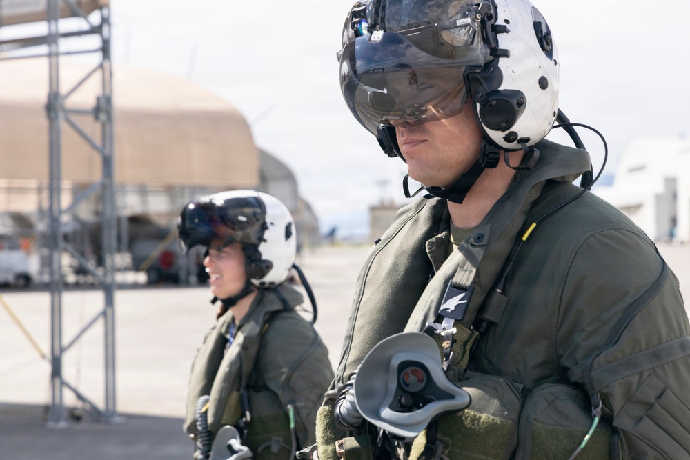 Two military aviators in full flight gear are standing on an airfield tarmac. Both are wearing white flight helmets with visors, olive drab flight suits, and communications equipment. A hardened aircraft shelter and ground support structures are visible in the background.