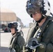 Two military aviators in full flight gear are standing on an airfield tarmac. Both are wearing white flight helmets with visors, olive drab flight suits, and communications equipment. A hardened aircraft shelter and ground support structures are visible in the background.