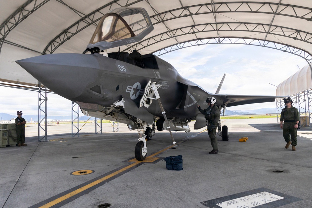A U.S. military F-35B stealth fighter jet, marked with the number '05' and a U.S. Navy/Marine Corps roundel, is parked under a large arched hangar shelter on a flight line. Several personnel in flight suits and helmets are conducting pre-flight or post-flight inspections around the aircraft. The cockpit canopy is open, and the landing gear is fully deployed.
