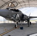 A U.S. military F-35B stealth fighter jet, marked with the number '05' and a U.S. Navy/Marine Corps roundel, is parked under a large arched hangar shelter on a flight line. Several personnel in flight suits and helmets are conducting pre-flight or post-flight inspections around the aircraft. The cockpit canopy is open, and the landing gear is fully deployed.