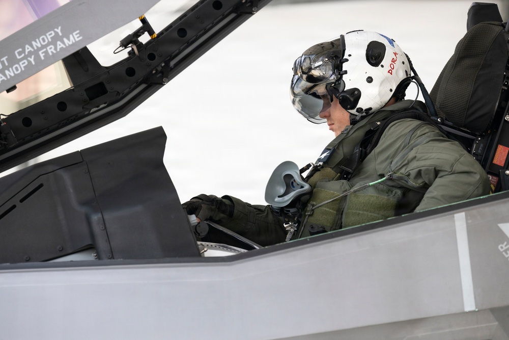 A military pilot is seated in the cockpit of a modern fighter jet, appearing to conduct pre-flight checks or instrument review. The pilot is wearing full flight gear including a white helmet with the call sign 'DORA' written in red letters, a visor, oxygen mask, flight suit, and gloves. The canopy frame is visible with partial text labeling including 'CANOPY FRAME'.