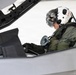 A military pilot is seated in the cockpit of a modern fighter jet, appearing to conduct pre-flight checks or instrument review. The pilot is wearing full flight gear including a white helmet with the call sign 'DORA' written in red letters, a visor, oxygen mask, flight suit, and gloves. The canopy frame is visible with partial text labeling including 'CANOPY FRAME'.