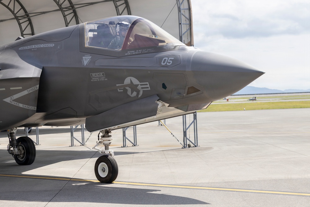 A U.S. military F-35 stealth fighter jet, marked with tail number '05', is parked on a tarmac in front of a hangar structure. A pilot is visible in the cockpit, suggesting pre-flight or post-flight operations. The aircraft displays U.S. Air Force/Marine Corps roundel insignia and multiple warning labels on its fuselage.