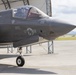 A U.S. military F-35 stealth fighter jet, marked with tail number '05', is parked on a tarmac in front of a hangar structure. A pilot is visible in the cockpit, suggesting pre-flight or post-flight operations. The aircraft displays U.S. Air Force/Marine Corps roundel insignia and multiple warning labels on its fuselage.