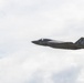 A single military jet aircraft is captured in flight against an overcast, cloudy sky. The aircraft displays U.S. Marine Corps markings and features a distinctive checkered tail fin design. The aircraft appears to be in level flight, photographed from a slightly low angle.