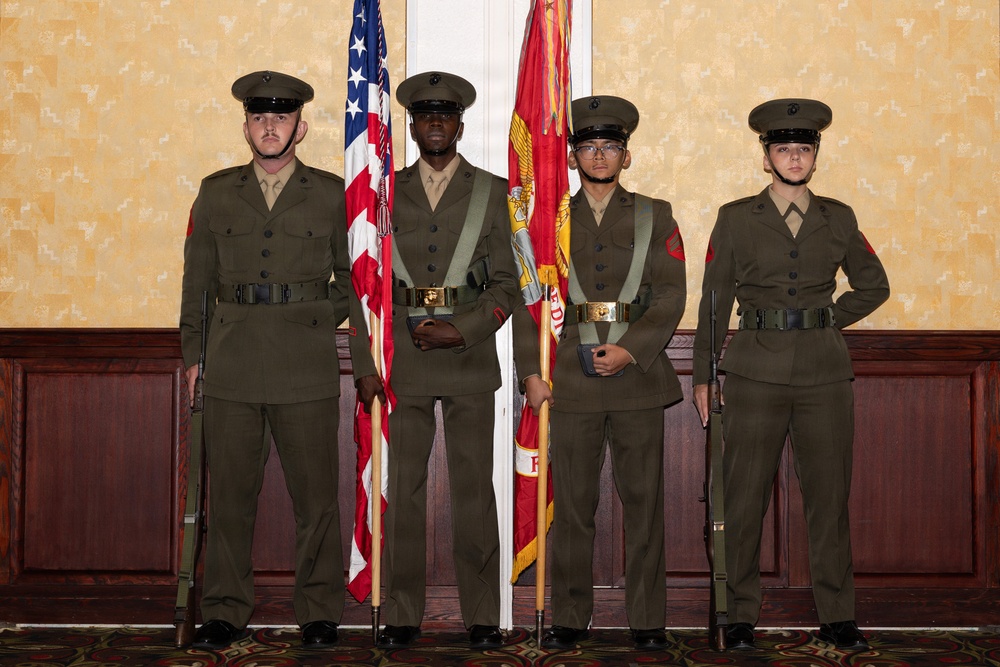 Four individuals in U.S. Marine Corps dress uniforms stand at attention in a color guard formation inside what appears to be a formal venue or banquet hall. Two members in the center are holding flags — the U.S. national flag and the U.S. Marine Corps flag — while the two on the flanks carry rifles at their sides. The formation is consistent with a ceremonial color guard presentation.