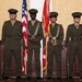 Four individuals in U.S. Marine Corps dress uniforms stand at attention in a color guard formation inside what appears to be a formal venue or banquet hall. Two members in the center are holding flags — the U.S. national flag and the U.S. Marine Corps flag — while the two on the flanks carry rifles at their sides. The formation is consistent with a ceremonial color guard presentation.