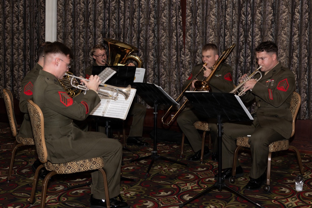 Four U.S. Marine Corps musicians in dress green uniforms are performing as a brass quintet in what appears to be a formal indoor venue. The musicians are seated and playing from sheet music on stands, with instruments including trumpets, a trombone, and a tuba/euphonium. The setting features ornate patterned curtains and carpeting consistent with a banquet hall or formal event space.
