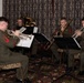 Four U.S. Marine Corps musicians in dress green uniforms are performing as a brass quintet in what appears to be a formal indoor venue. The musicians are seated and playing from sheet music on stands, with instruments including trumpets, a trombone, and a tuba/euphonium. The setting features ornate patterned curtains and carpeting consistent with a banquet hall or formal event space.