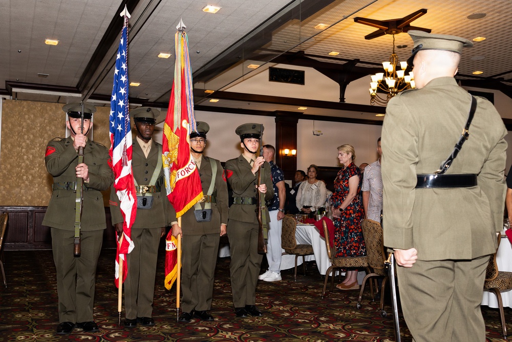 A U.S. Marine Corps Color Guard ceremony is taking place inside what appears to be a banquet hall or dining facility. Four Marines in dress uniforms are presenting the colors, including the American flag and a red Marine Corps unit guidon, while an officer faces them and civilian guests stand at attention in the background. The event appears to be a formal military dining-in or awards ceremony.