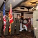 A U.S. Marine Corps Color Guard ceremony is taking place inside what appears to be a banquet hall or dining facility. Four Marines in dress uniforms are presenting the colors, including the American flag and a red Marine Corps unit guidon, while an officer faces them and civilian guests stand at attention in the background. The event appears to be a formal military dining-in or awards ceremony.
