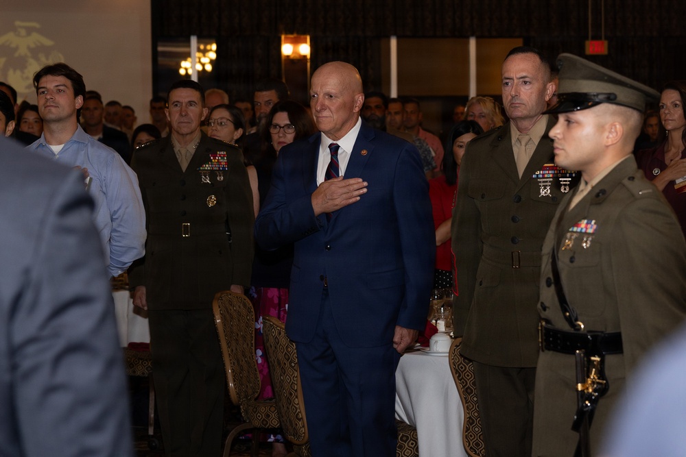 A formal military or civic ceremony is taking place in a banquet hall setting. A civilian man in a blue suit stands at attention with his hand over his heart, flanked by uniformed U.S. Marine Corps officers wearing service uniforms with medal ribbons. A large crowd of attendees stands in the background, suggesting a pledge of allegiance or national anthem moment.