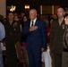 A formal military or civic ceremony is taking place in a banquet hall setting. A civilian man in a blue suit stands at attention with his hand over his heart, flanked by uniformed U.S. Marine Corps officers wearing service uniforms with medal ribbons. A large crowd of attendees stands in the background, suggesting a pledge of allegiance or national anthem moment.