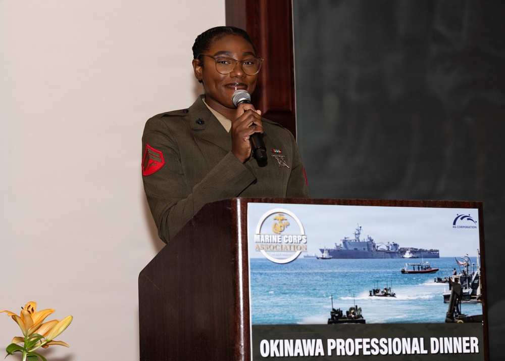 A U.S. Marine in service uniform is speaking at a podium during the Marine Corps Association Okinawa Professional Dinner. The speaker appears to be a non-commissioned officer, identifiable by the red chevron patch on the left sleeve of an olive green service uniform. The event signage on the podium displays the Marine Corps Association logo and a naval amphibious operations image.