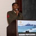 A U.S. Marine in service uniform is speaking at a podium during the Marine Corps Association Okinawa Professional Dinner. The speaker appears to be a non-commissioned officer, identifiable by the red chevron patch on the left sleeve of an olive green service uniform. The event signage on the podium displays the Marine Corps Association logo and a naval amphibious operations image.