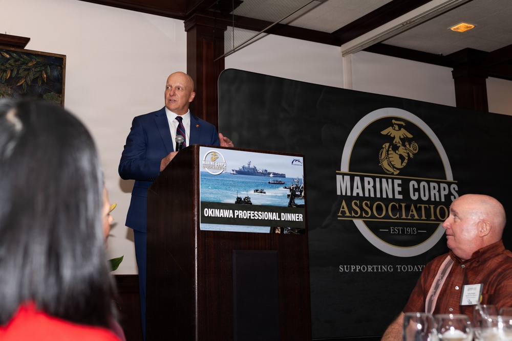A man in a blue suit is delivering a speech at a podium during the Marine Corps Association Okinawa Professional Dinner. A presentation screen behind the podium displays the event title 'Okinawa Professional Dinner' with a military amphibious landing image. The event banner prominently features the Marine Corps Association logo, established in 1913.