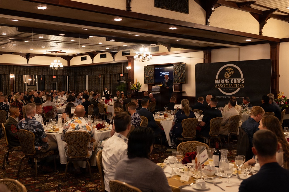 A formal banquet or dinner event hosted by the Marine Corps Association (Est. 1913) is taking place in an elegantly decorated ballroom. A speaker stands at a podium on a raised stage area, addressing a large audience seated at round tables with white tablecloths. The event appears well-attended with military and civilian guests dressed in a mix of formal attire and military uniforms.