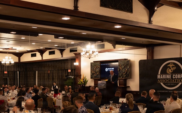 A formal banquet or dinner event hosted by the Marine Corps Association (Est. 1913) is taking place in an elegantly decorated ballroom. A speaker stands at a podium on a raised stage area, addressing a large audience seated at round tables with white tablecloths. The event appears well-attended with military and civilian guests dressed in a mix of formal attire and military uniforms.