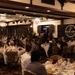 A formal banquet or dinner event hosted by the Marine Corps Association (Est. 1913) is taking place in an elegantly decorated ballroom. A speaker stands at a podium on a raised stage area, addressing a large audience seated at round tables with white tablecloths. The event appears well-attended with military and civilian guests dressed in a mix of formal attire and military uniforms.