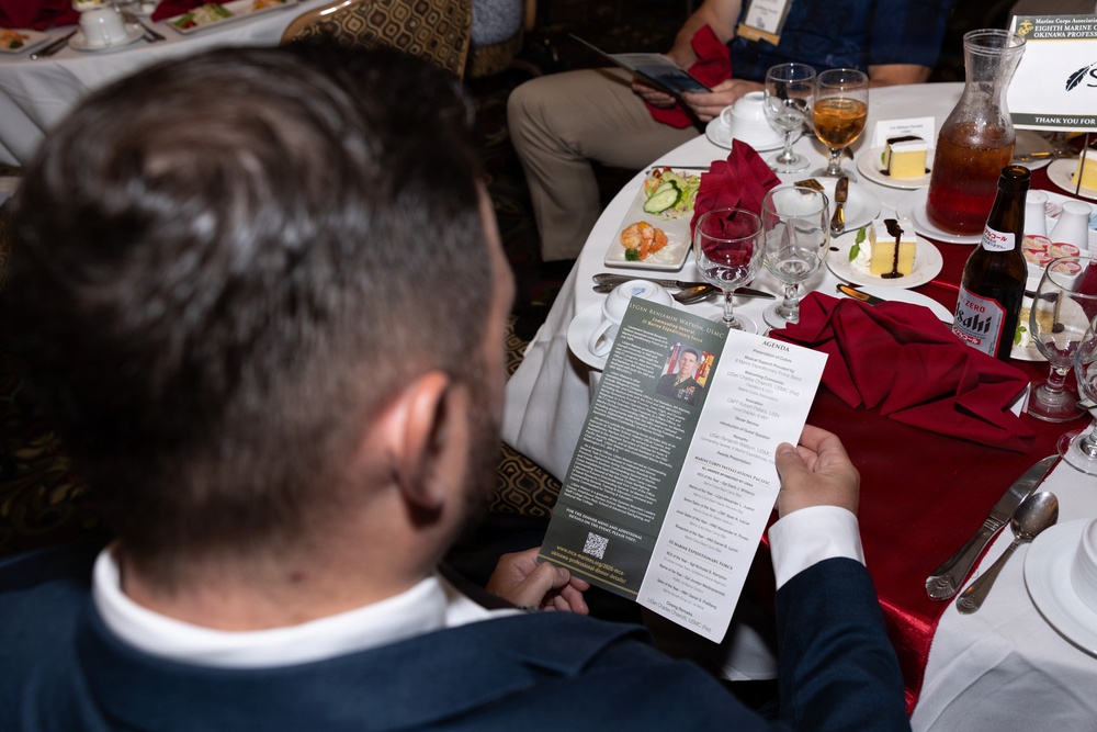 A man in a dark suit is seated at a formal banquet table, reading a printed program or brochure that appears to be for a Marine Corps Association event. The table is set with red napkins, wine glasses, dessert plates, and Asahi beer bottles, suggesting a formal military dinner or awards ceremony. A sign in the background references 'Eighth Marines Okinawa' and a 'Marine Corps Association' event.