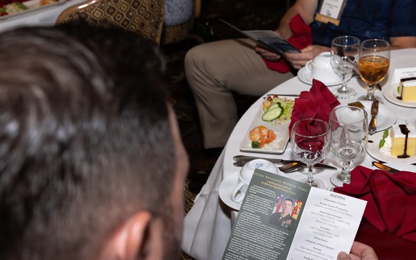 A man in a dark suit is seated at a formal banquet table, reading a printed program or brochure that appears to be for a Marine Corps Association event. The table is set with red napkins, wine glasses, dessert plates, and Asahi beer bottles, suggesting a formal military dinner or awards ceremony. A sign in the background references 'Eighth Marines Okinawa' and a 'Marine Corps Association' event.